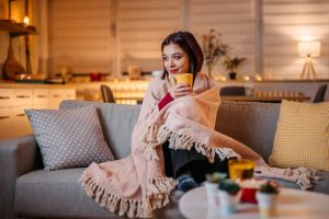 Beautiful woman with coffee cosy up to a wrapped in blanket on sofa at home.
