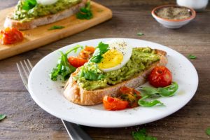 Avocado toast with roasted tomatoes and eggs, on a white plate on a rustic wooden table. Served with fresh arugula and sprinkled with parsley and black pepper. Stock photo.