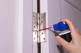 Close up of man hand using spray can to grease metal hinge on white door.