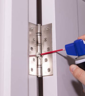 Close up of man hand using spray can to grease metal hinge on white door.
